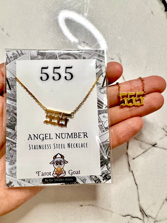 A gold-toned stainless steel necklace with an '555 Angel Number' pendant is displayed on its branded retail card, held by a hand over a white marble surface.
