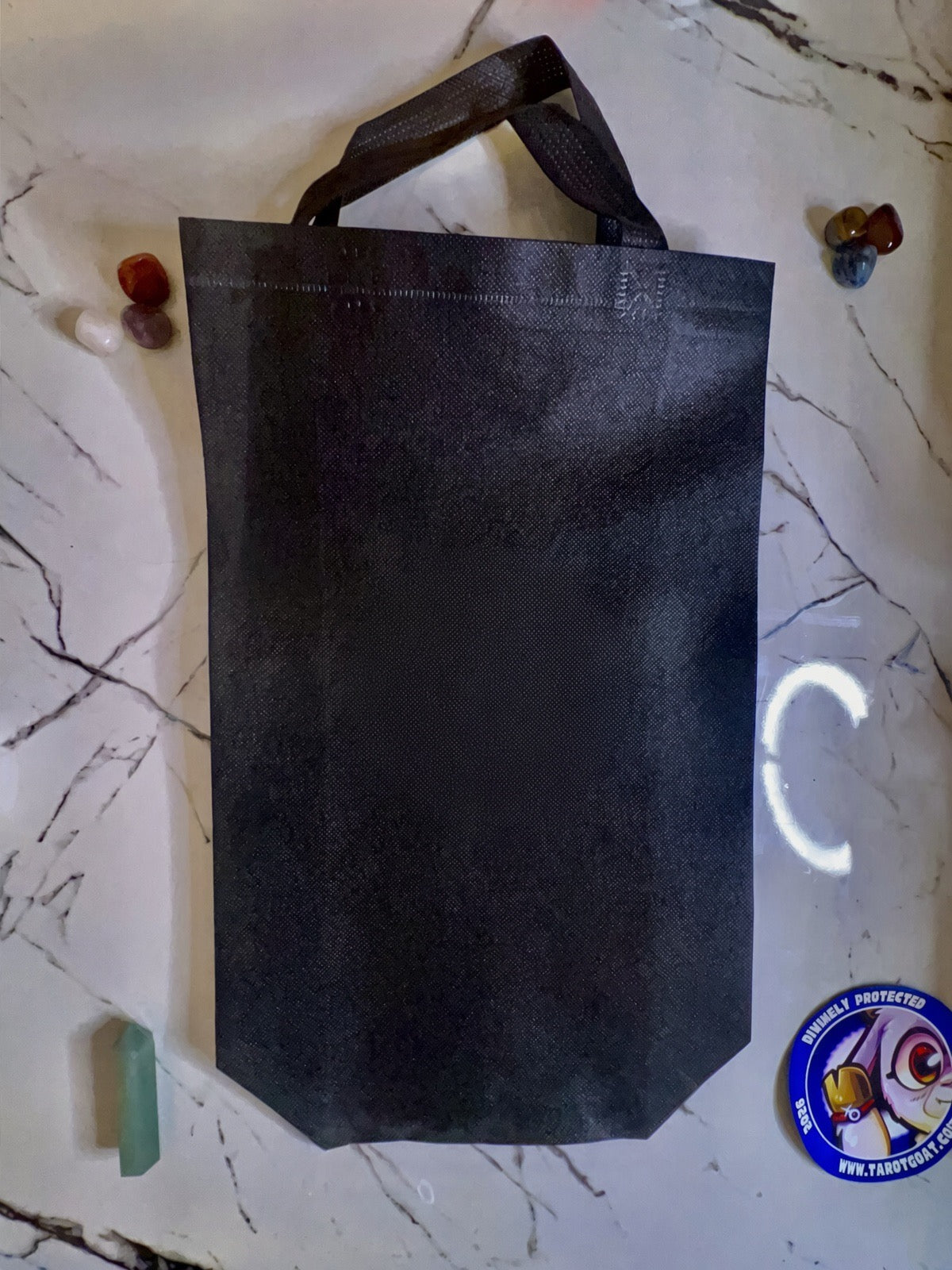 A black reusable tote bag with two handles, laid flat on a white marble-patterned surface with small decorative stones around it.