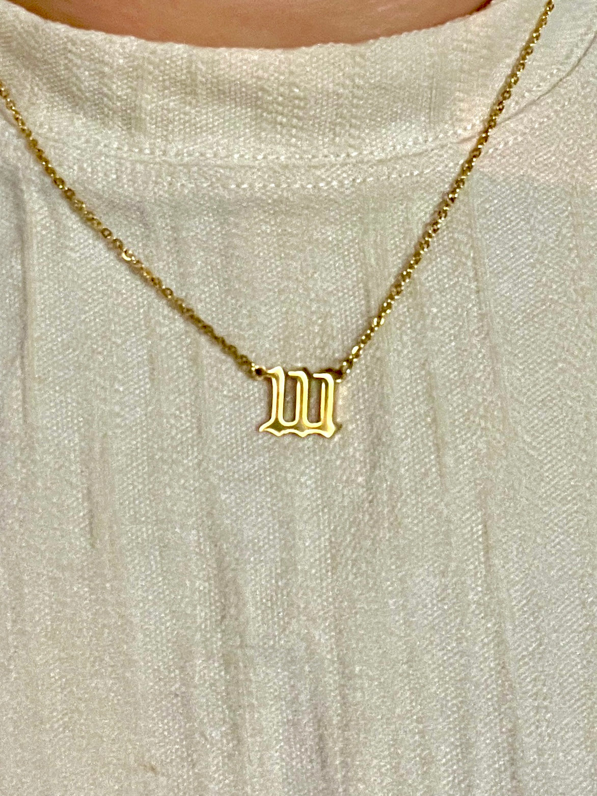 A close-up of a gold necklace with with the number 111  pendant, worn on a person's neck over a textured cream-colored top.