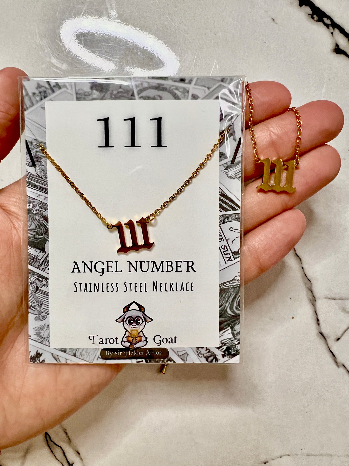 A hand holds a packaged stainless steel necklace with a Roman numeral III pendant. The card reads '111 ANGEL NUMBER STAINLESS STEEL NECKLACE' and shows the brand Tarot Goat.