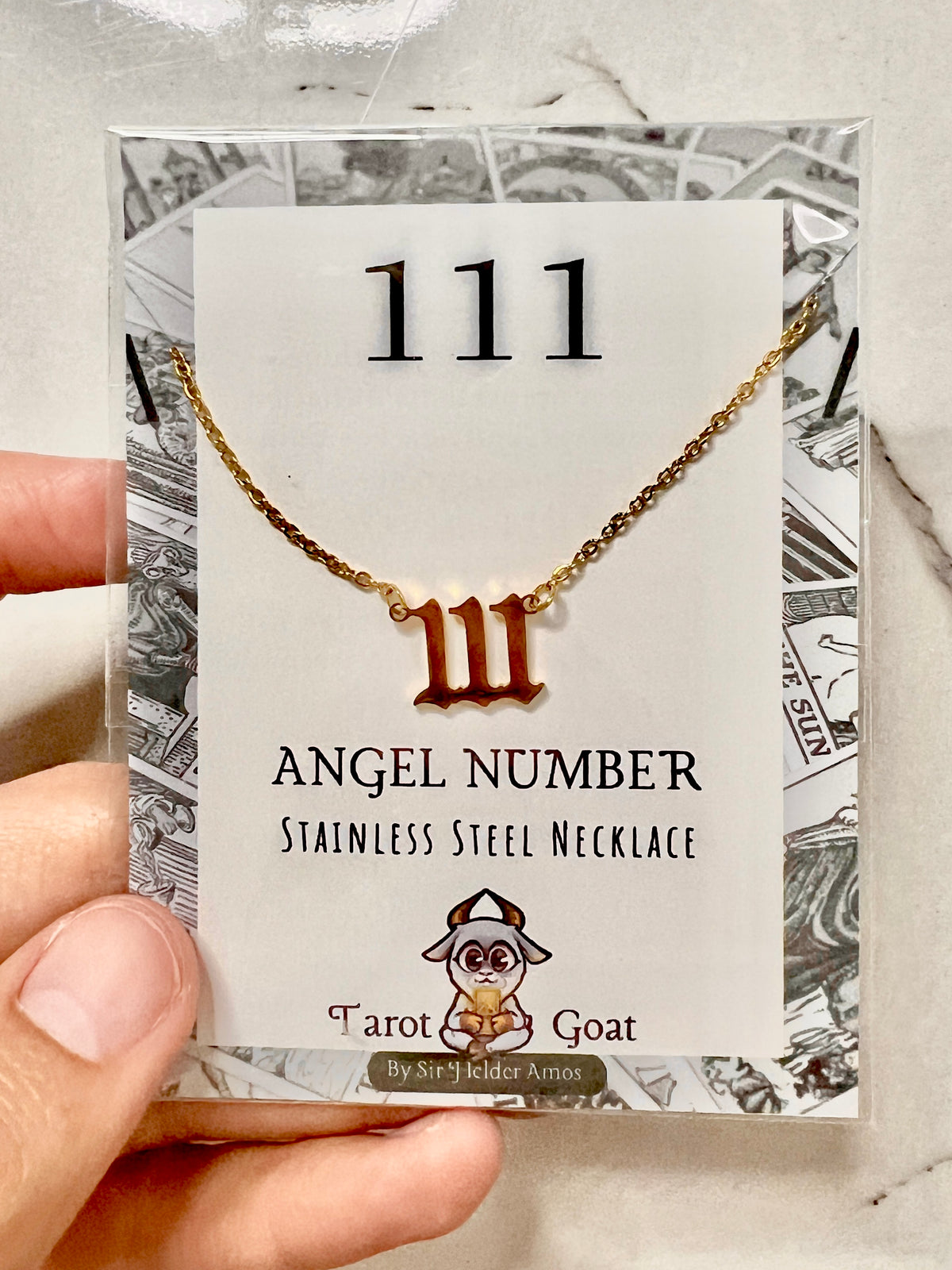 A gold-tone stainless steel necklace with a Roman numeral III pendant, displayed on a branded backing card held by a hand. The card reads '111 ANGEL NUMBER STAINLESS STEEL NECKLACE' and shows the brand 'Tarot Goat' with a goat illustration.