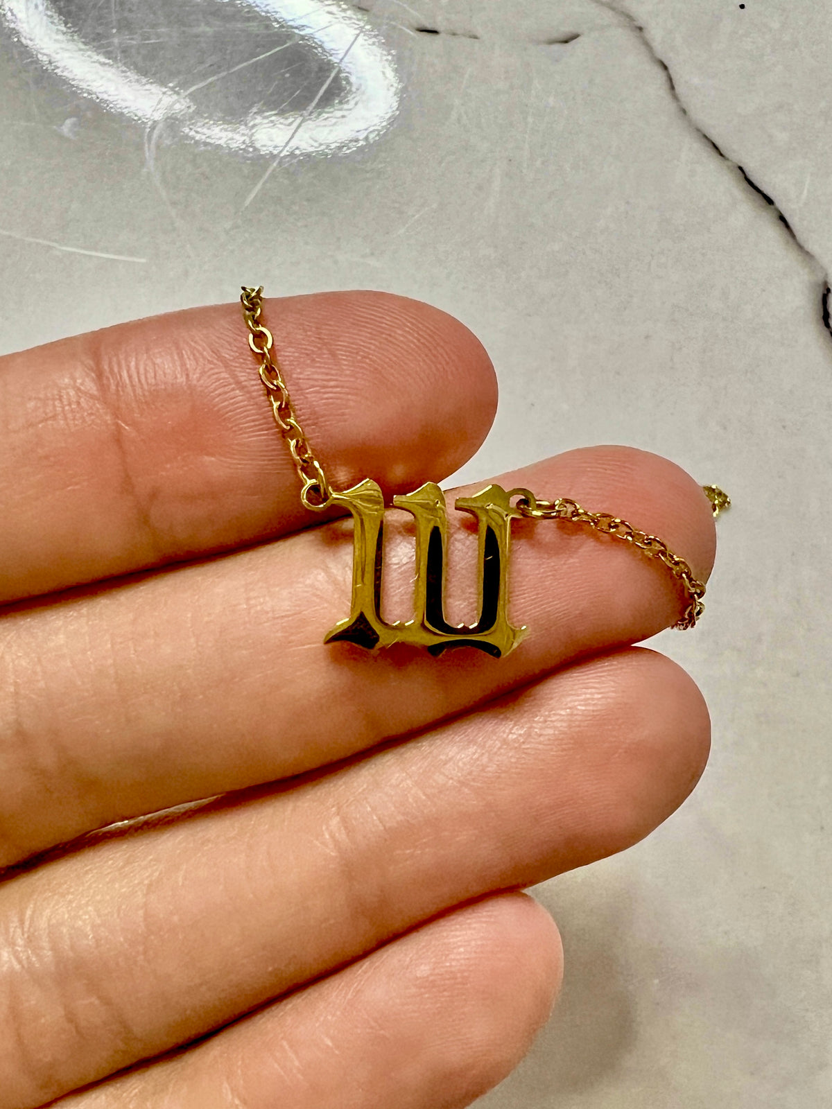 A gold-toned pendant necklace shaped like the Roman numeral 'III' held on a person's fingers against a light marble-like background.