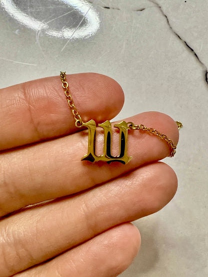 A gold-toned pendant necklace shaped like the Roman numeral 'III' held on a person's fingers against a light marble-like background.