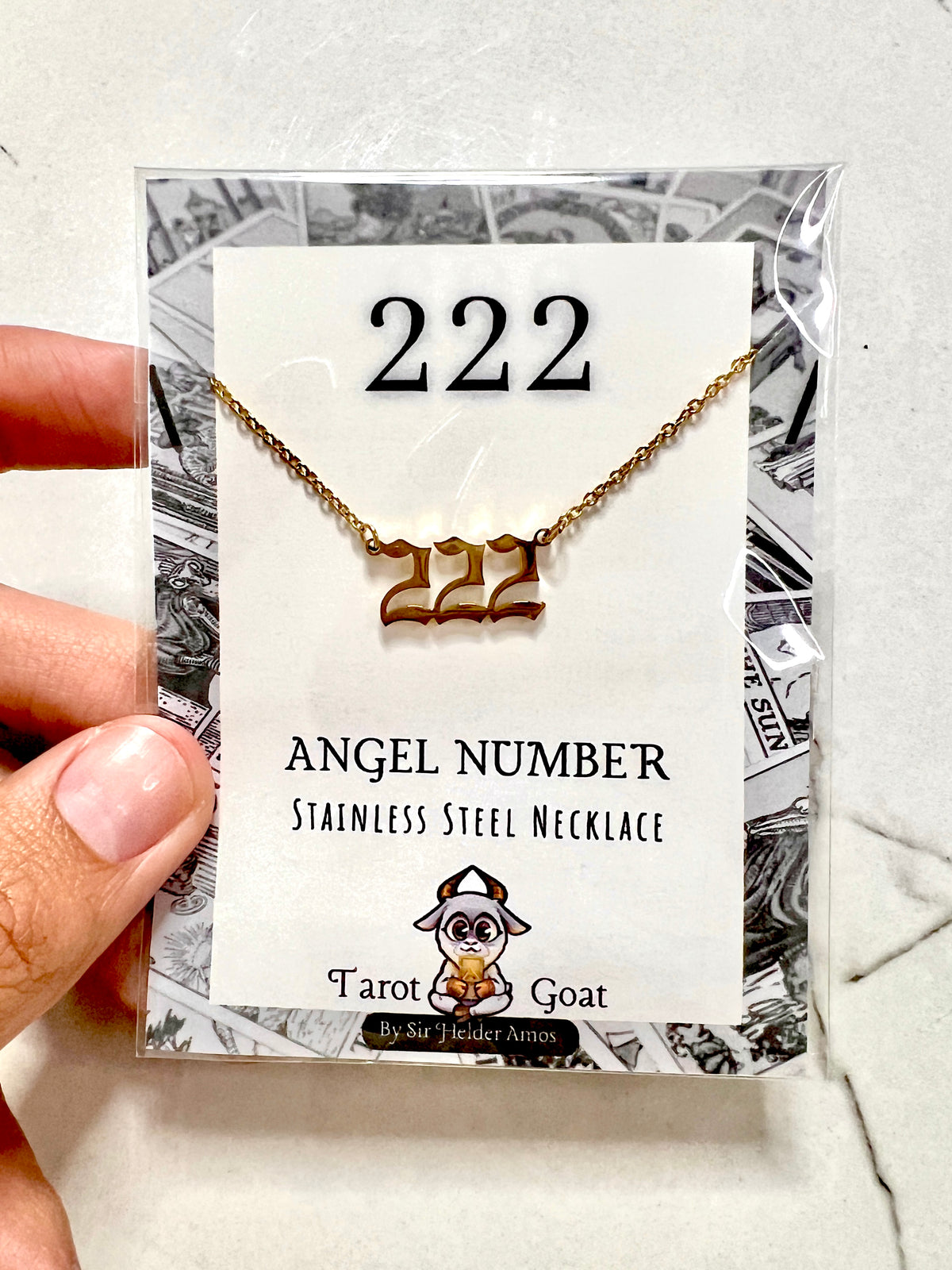 A stainless steel necklace with the number '222' pendant, packaged on a branded display card and held by a hand. The card reads '222 ANGEL NUMBER STAINLESS STEEL NECKLACE' and shows the Tarot Goat brand.