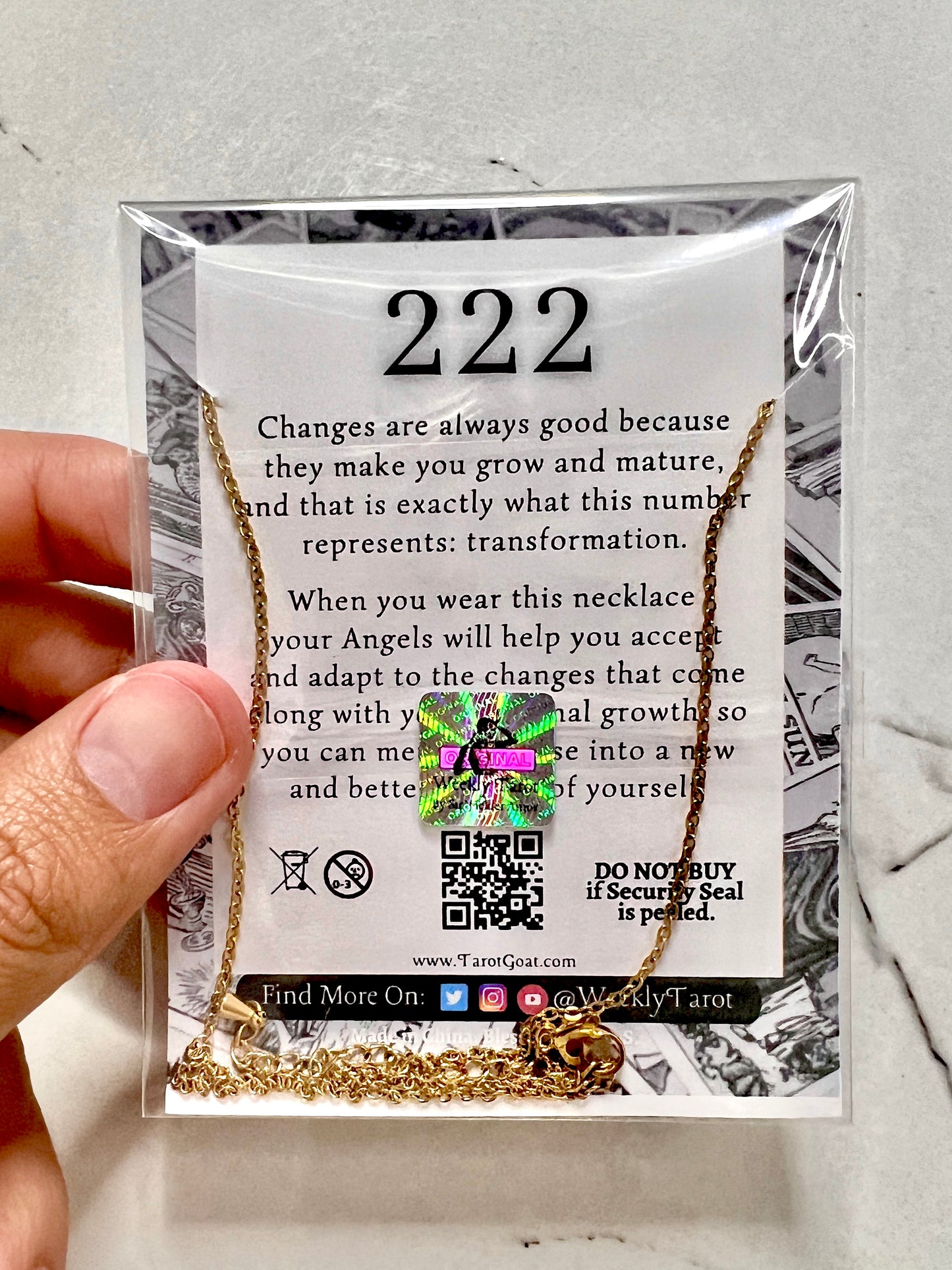 A gold-toned 222 necklace displayed inside a clear plastic sleeve on a printed card. A hand is holding the left side of the package, which features the number 222 and inspirational text about transformation.