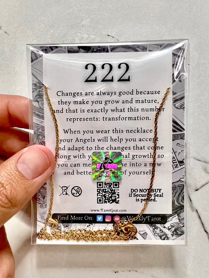 A gold-toned 222 necklace displayed inside a clear plastic sleeve on a printed card. A hand is holding the left side of the package, which features the number 222 and inspirational text about transformation.
