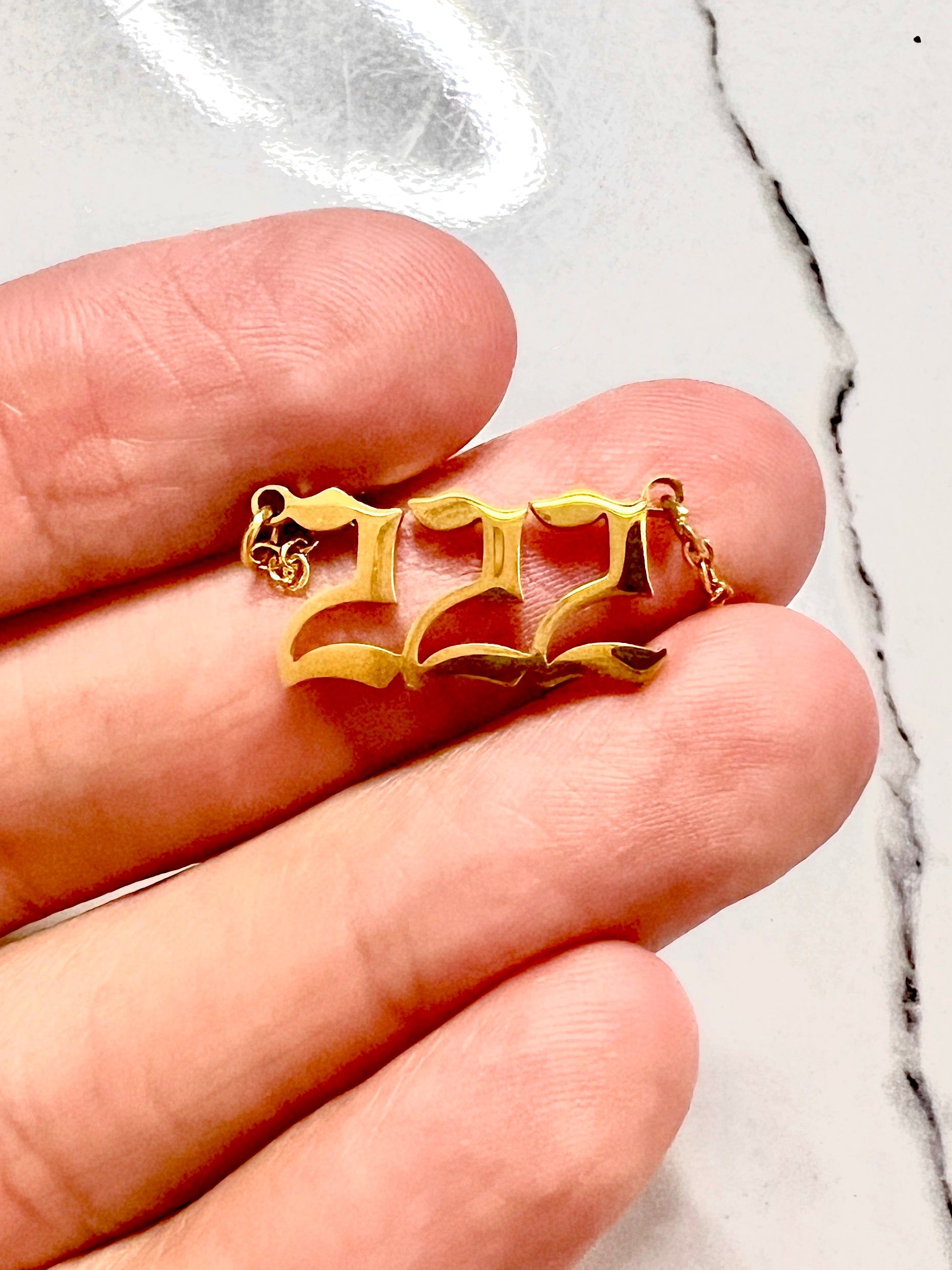 A gold pendant with the angel number 222, resting on a person's fingers against a white marble background.