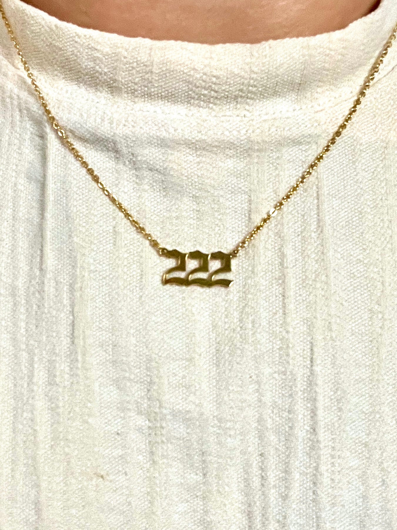 A gold chain necklace with a '222' number pendant worn by a person wearing a textured off-white linen top.