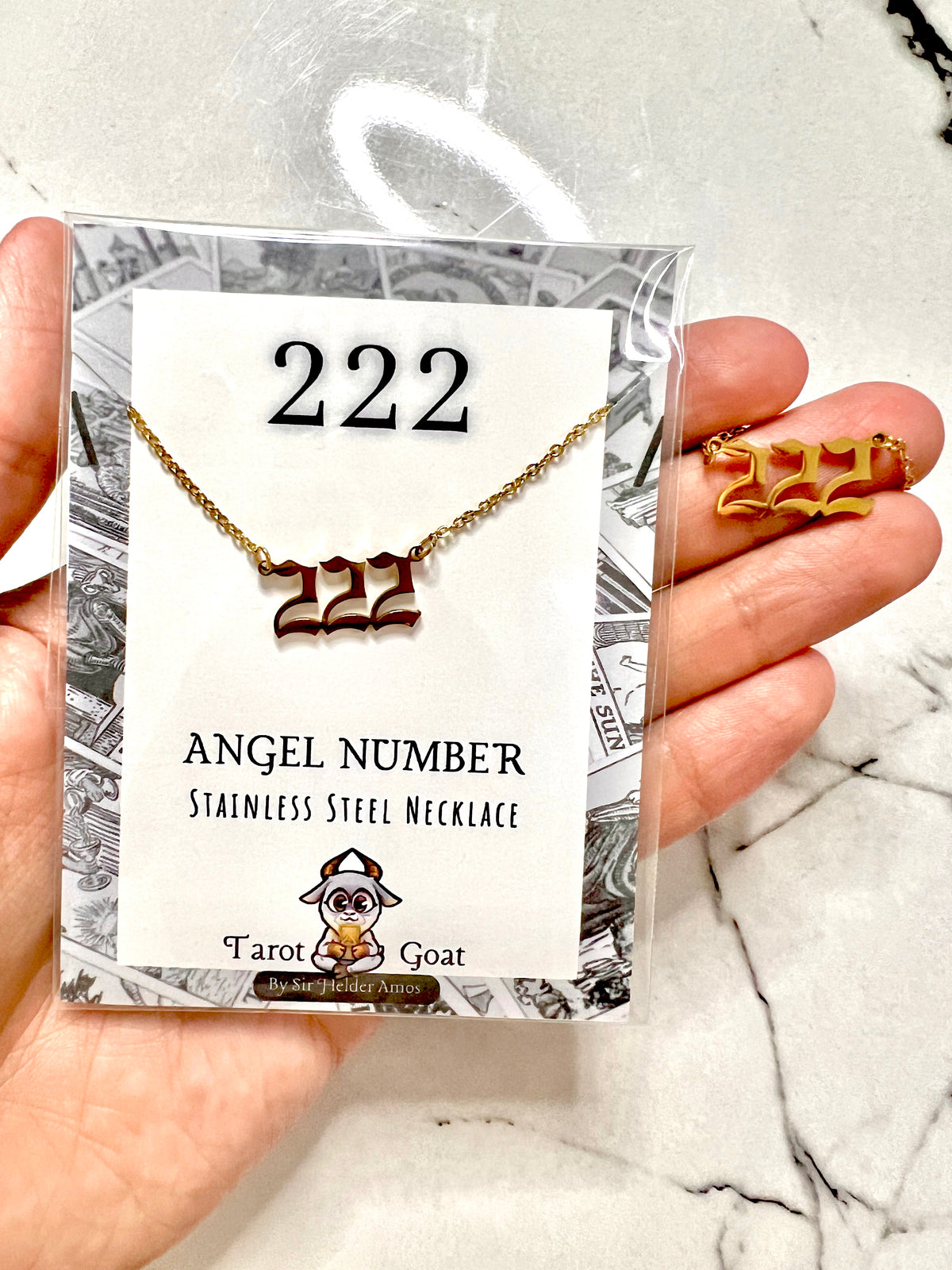 A hand holds a packaged stainless steel necklace and matching stud earrings featuring the number 222, with the card reading '222 ANGEL NUMBER STAINLESS STEEL NECKLACE' and the brand 'Tarot Goat'.