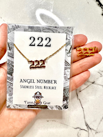 A hand holds a packaged stainless steel necklace and matching stud earrings featuring the number 222, with the card reading '222 ANGEL NUMBER STAINLESS STEEL NECKLACE' and the brand 'Tarot Goat'.