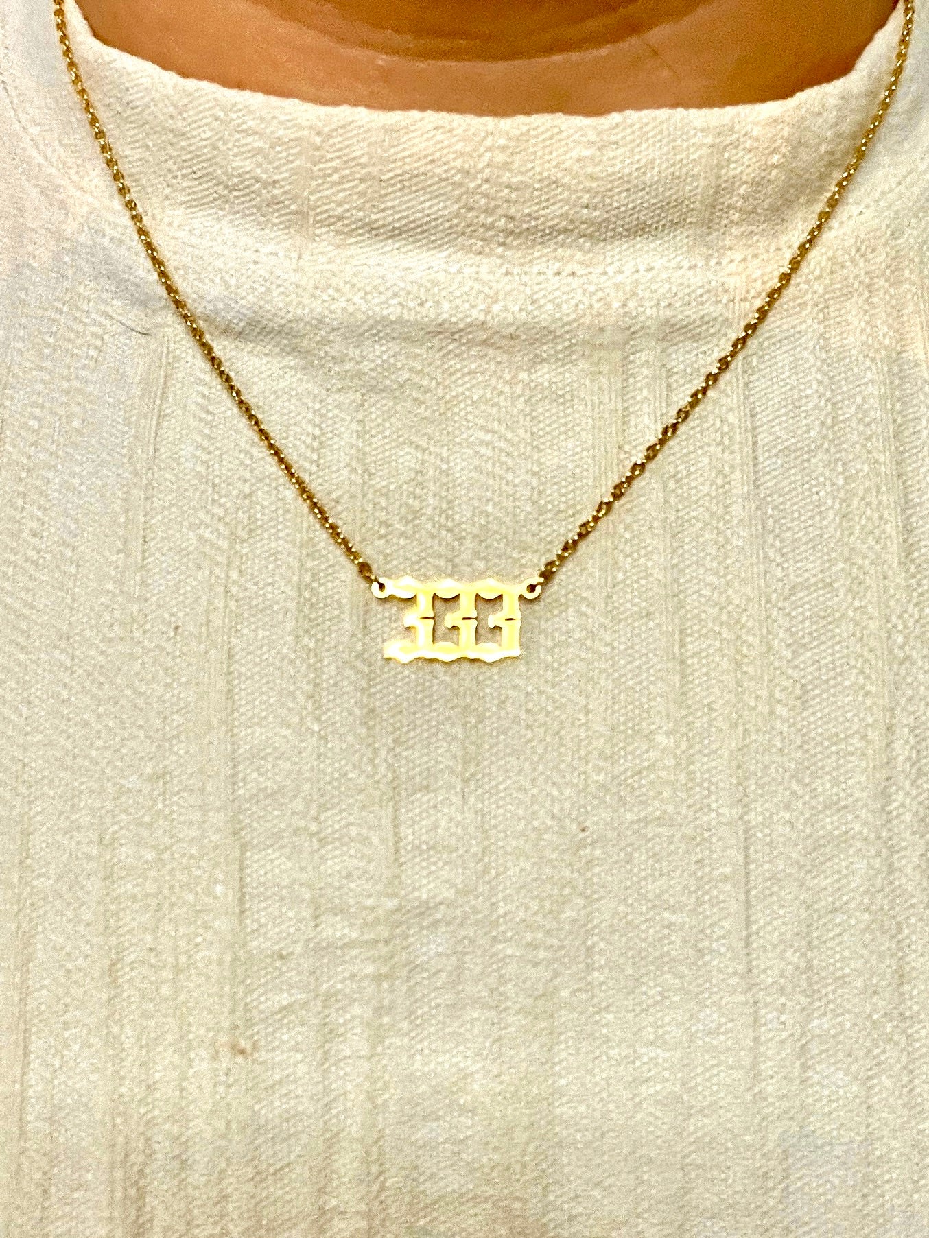 A gold chain necklace with a 333 number pendant, worn on a person's neck over a cream-colored knit top.