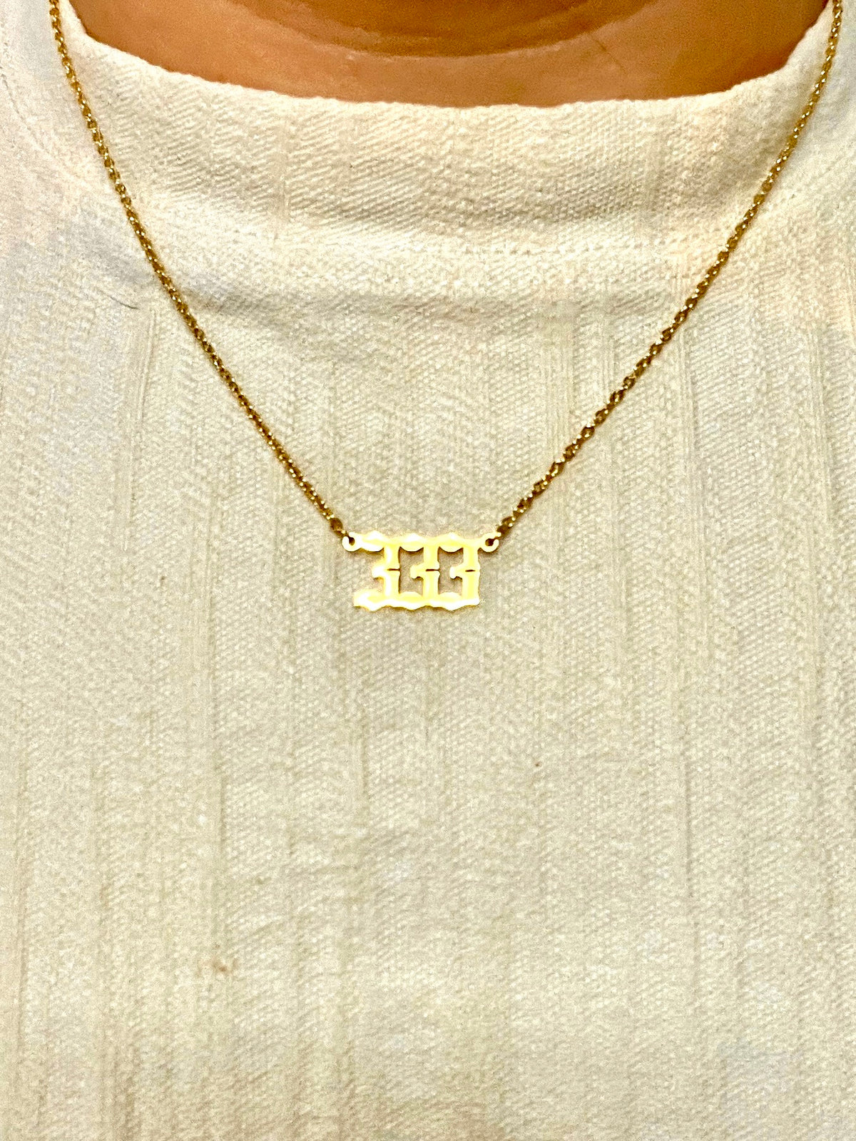 A gold chain necklace with a 333 number pendant, worn on a person's neck over a cream-colored knit top.