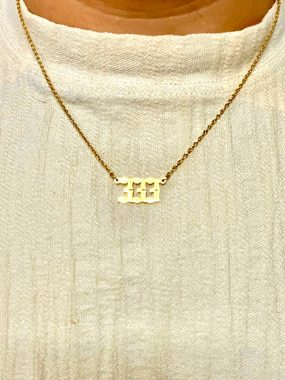 A gold chain necklace with a 333 number pendant, worn on a person's neck over a cream-colored knit top.