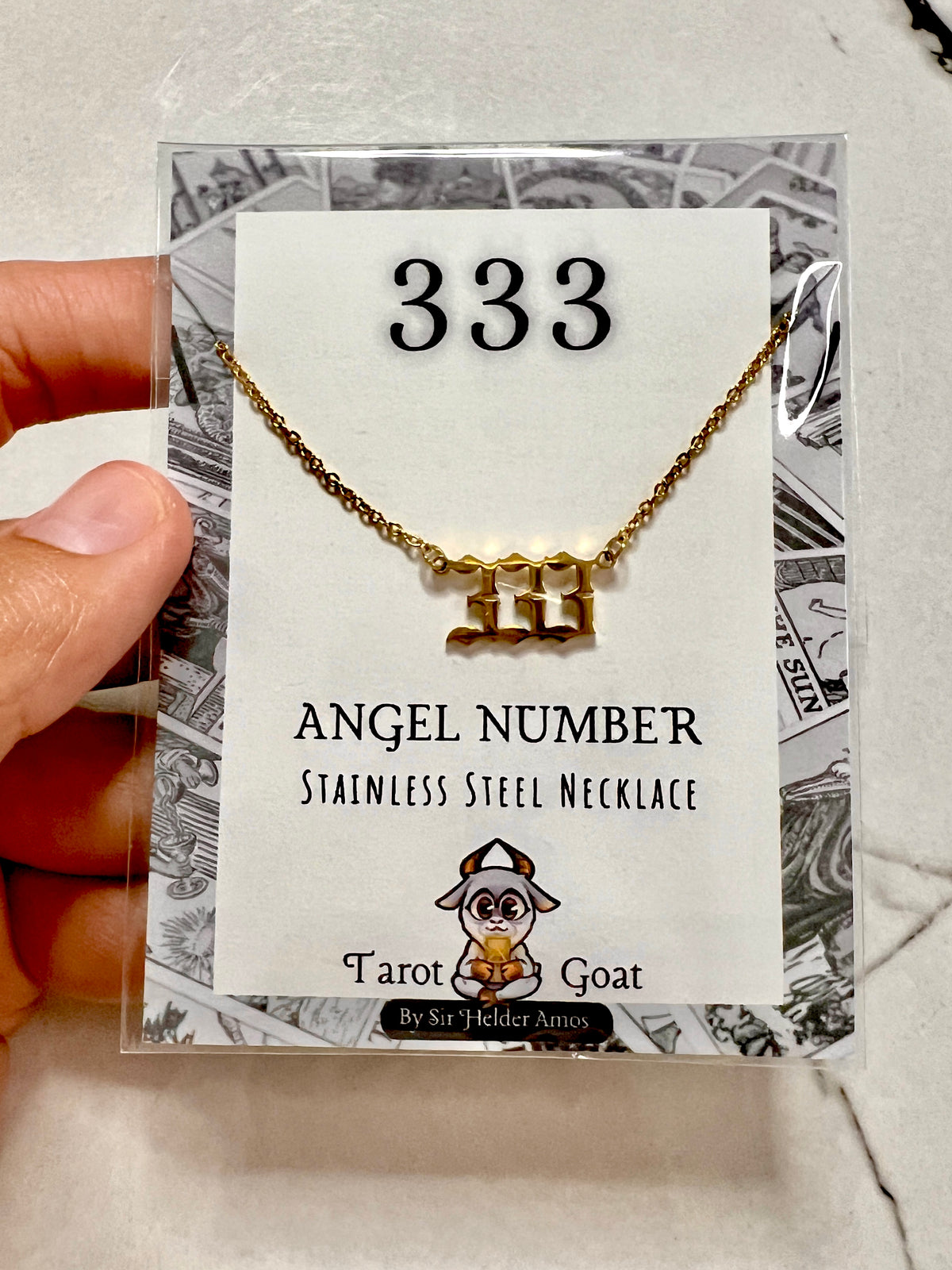 A hand holds a clear plastic package containing a gold-tone stainless steel necklace with a stylized '333' pendant, displayed on a white card labeled '333' and 'Angel Number Stainless Steel Necklace' with the Tarot Goat logo.