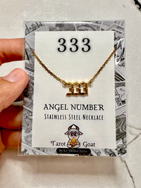 A hand holds a clear plastic package containing a gold-tone stainless steel necklace with a stylized '333' pendant, displayed on a white card labeled '333' and 'Angel Number Stainless Steel Necklace' with the Tarot Goat logo.