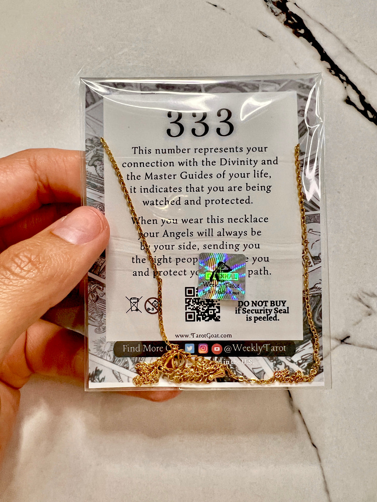 A hand holding a clear plastic package containing a gold-toned necklace with a white card that reads '333' and includes a security seal. The background is a light marble surface.