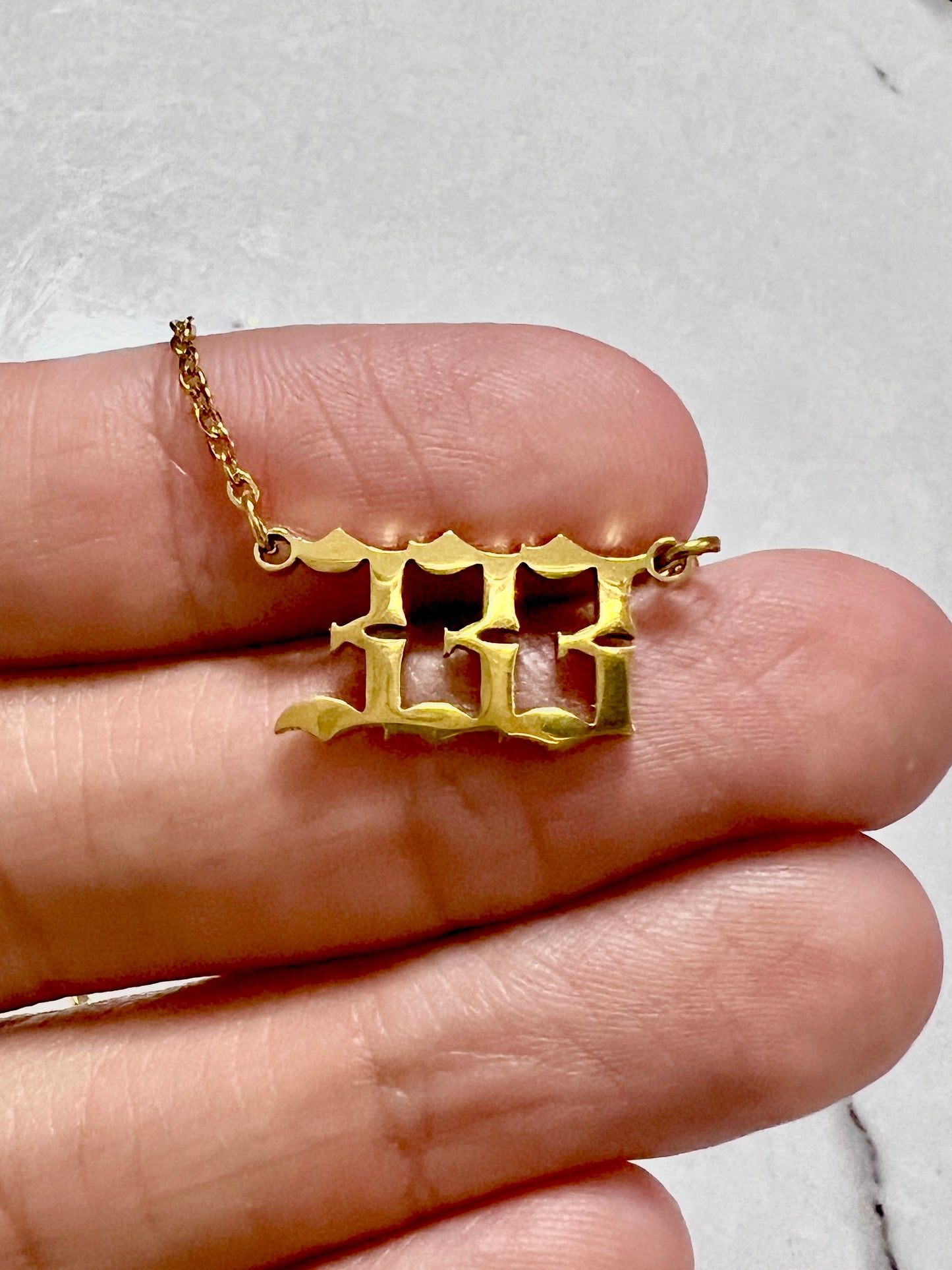 A gold-toned pendant necklace with a stylized 333 number design, shown resting on a person's fingers against a light gray marble background.