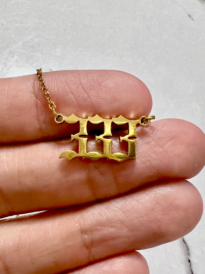 A gold-toned pendant necklace with a stylized 333 number design, shown resting on a person's fingers against a light gray marble background.