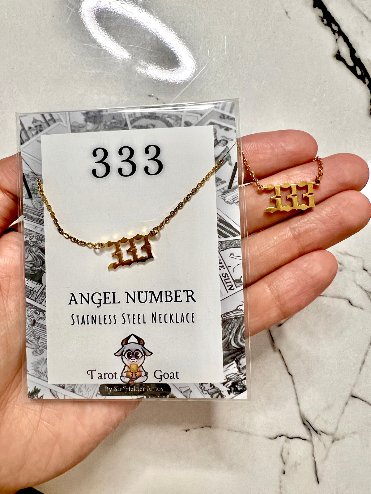 A hand holds a gold-tone stainless steel necklace with a triple-three symbol pendant, displayed on a branded backing card labeled '333 Angel Number Stainless Steel Necklace' by Tarot Goat, against a light marble surface.