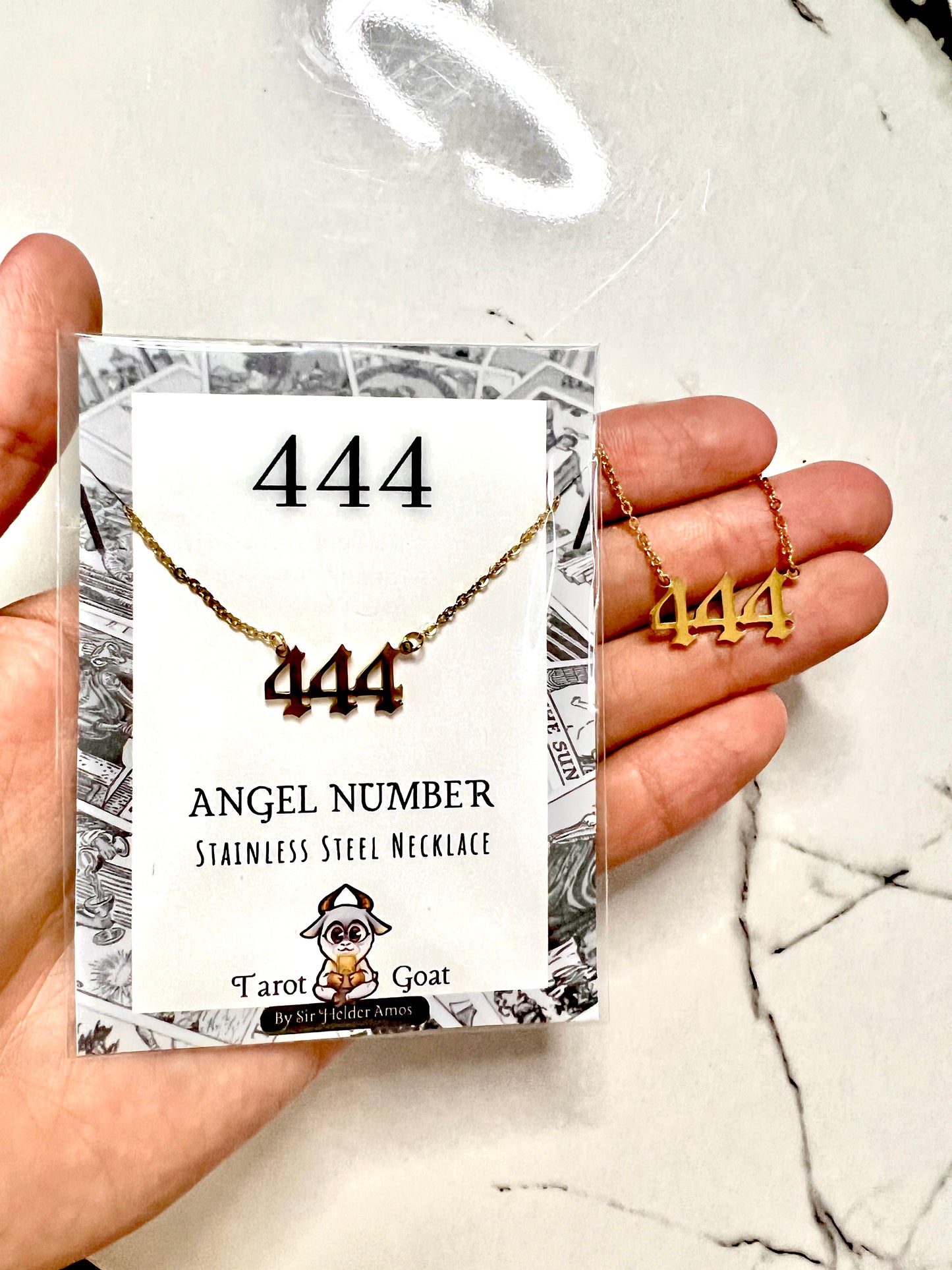 A hand holds a packaged stainless steel necklace with three '444' angel numbers on a card. The necklace is gold-toned, and the packaging reads '444 ANGEL NUMBER STAINLESS STEEL NECKLACE' with a 'Tarot to Treasure Goat' logo.