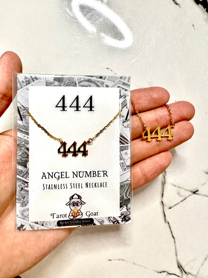 A hand holds a packaged stainless steel necklace with three '444' angel numbers on a card. The necklace is gold-toned, and the packaging reads '444 ANGEL NUMBER STAINLESS STEEL NECKLACE' with a 'Tarot to Treasure Goat' logo.