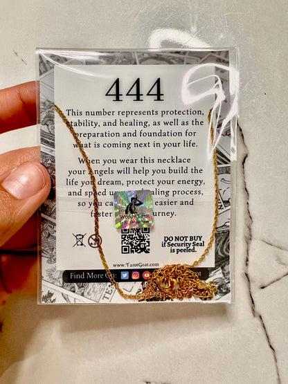 A hand holding a gold necklace in its original retail packaging. The packaging features a white card with black text detailing the meaning of the number 444, accompanied by a holographic security seal and a small illustration of a gold angel.