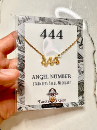 A hand holds a packaged gold necklace with a '444' pendant on a white display card. The card reads '444 ANGEL NUMBER STAINLESS STEEL NECKLACE' and shows the brand Tarot Goat