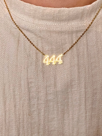 A close-up of a person wearing a gold chain necklace with a '444' pendant, styled over a textured cream-colored knit top.