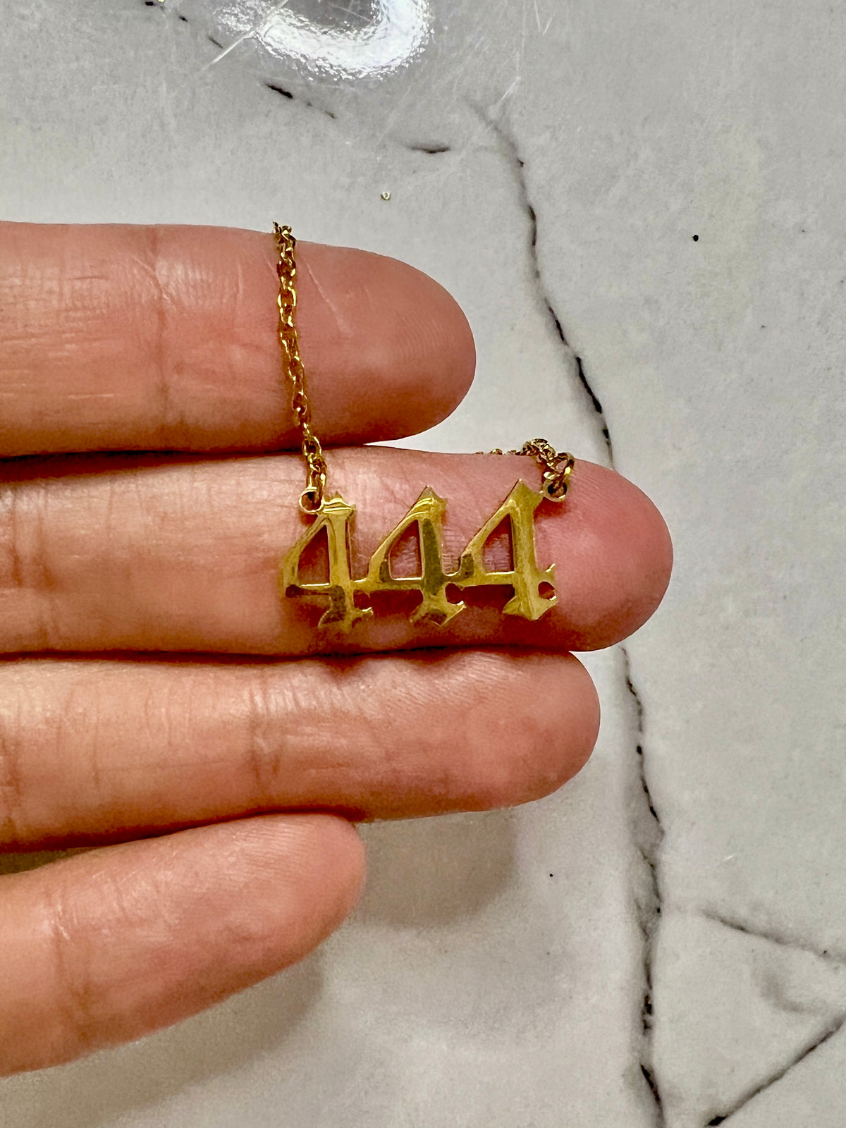 A gold necklace with a '444' pendant held on a person's fingers against a light marble background.