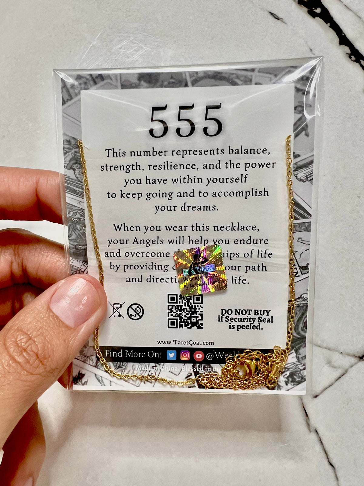 A hand holding a clear plastic package containing a gold-toned necklace with a small iridescent security seal and a white card printed with the number 555 and descriptive text, photographed over a light marble surface.