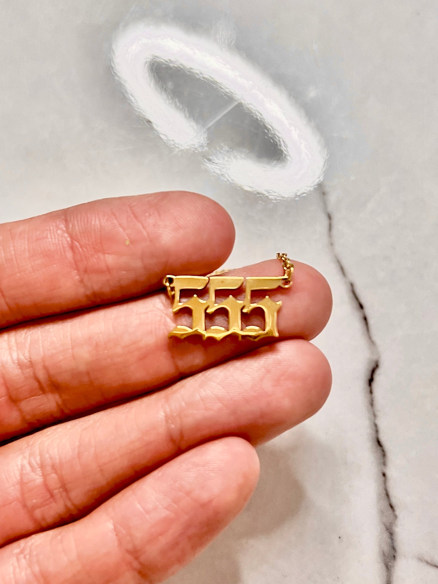 A gold-plated 555  number pendant charm held on a person's fingers against a light gray marble background for numerology