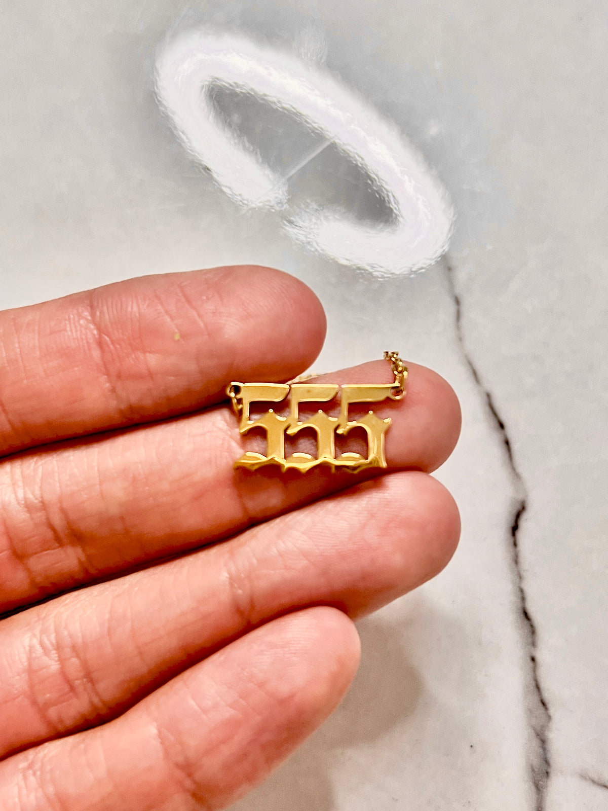 A gold-plated 555  number pendant charm held on a person's fingers against a light gray marble background for numerology