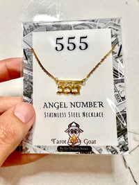 A hand holding a packaged stainless steel necklace with a gold-tone '555' angel number pendant on a branded display card.