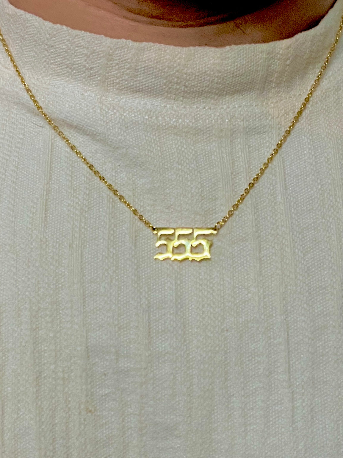 A close-up of a person wearing a delicate gold chain necklace with a small gold pendant shaped like the number 555, styled with a ribbed cream knit top.
