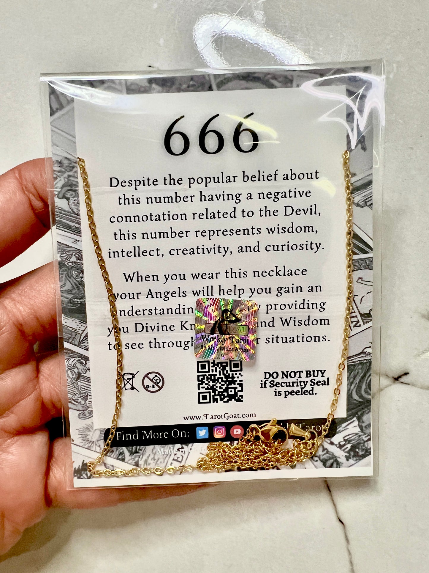A hand holds a clear plastic-wrapped jewelry card featuring a gold 666 necklace with a colorful crystal pendant. The card includes printed text about the number 666 and a QR code, photographed against a light stone surface.