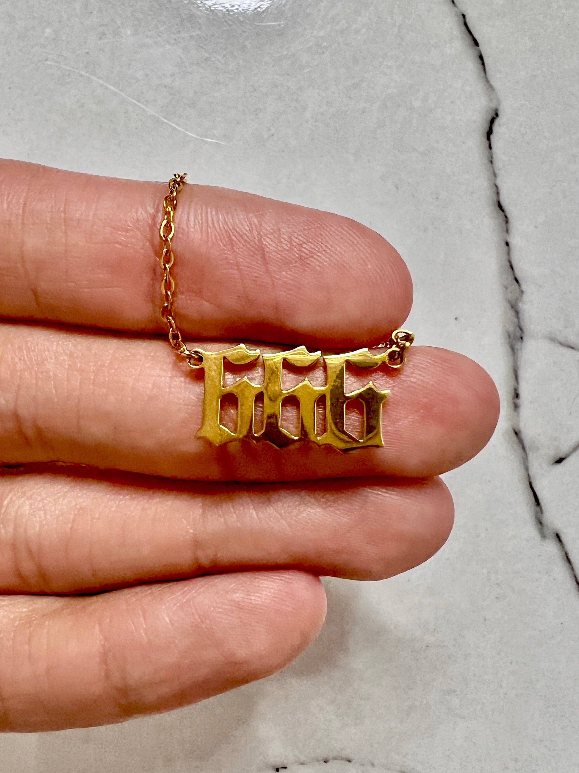 A gold necklace with the number 666  in a gothic font, held on a person's fingers against a light gray marble background.