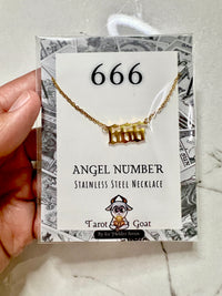 A hand holding a packaged stainless steel necklace with a gold-tone '666' angel number pendant on a white display card. The card reads '666 ANGEL NUMBER STAINLESS STEEL NECKLACE' and shows the Tarot Goat brand.