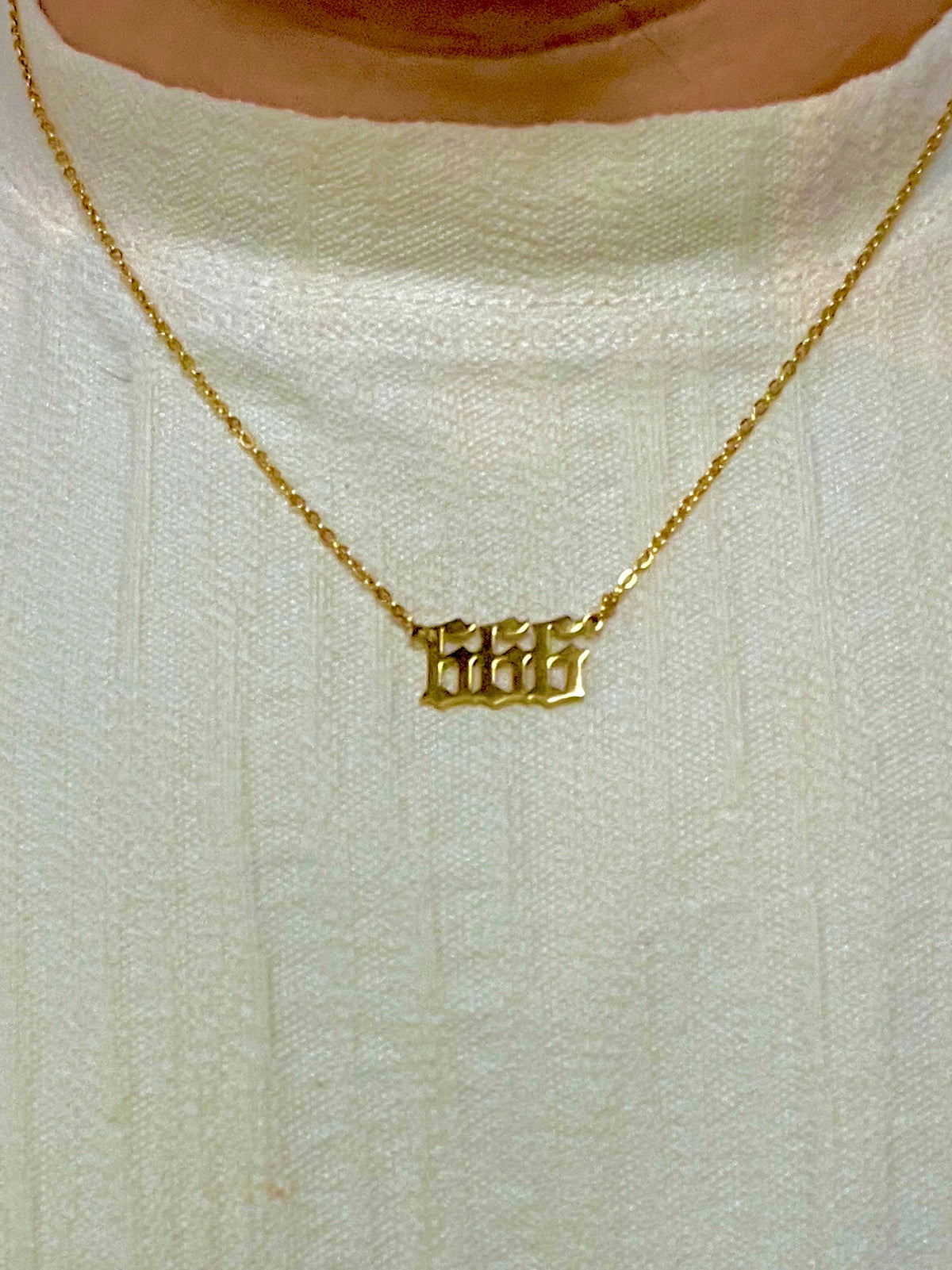 A close-up of a person wearing a delicate gold chain necklace with the angel number 666 in a gothic font, over a textured off-white knit top.