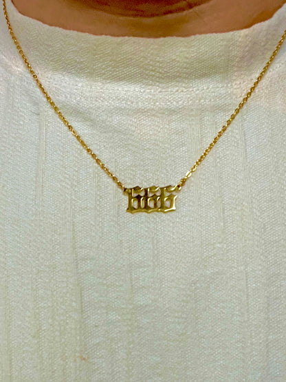 A close-up of a person wearing a delicate gold chain necklace with the angel number 666 in a gothic font, over a textured off-white knit top.