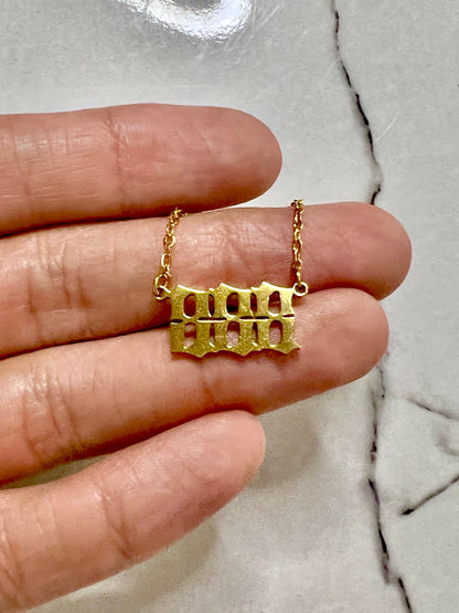 A gold-toned  888 necklace displayed on a person's fingers against a light marble background.