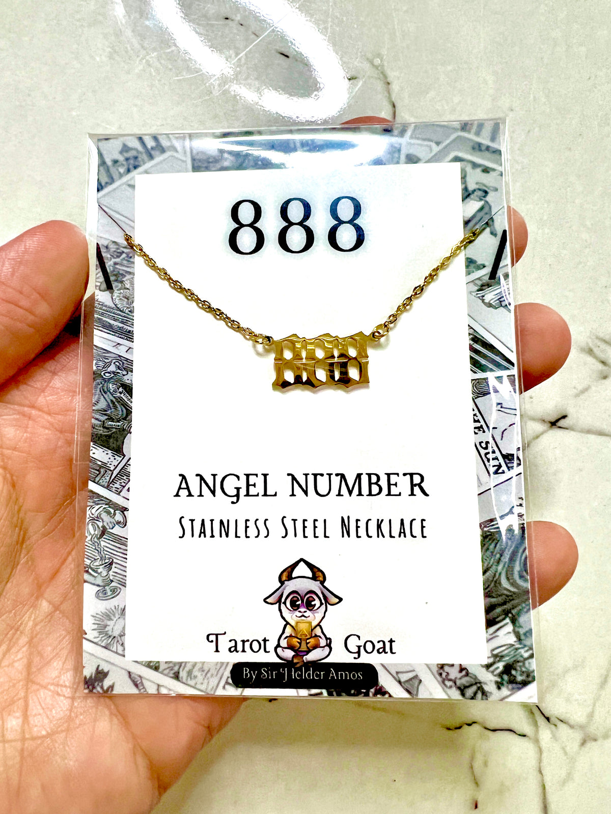 A hand holding a packaged gold-tone stainless steel 888 number necklace with a rectangular 'angel number' pendant. The necklace is displayed on a white card labeled '888' and 'Angel Number Stainless Steel Necklace' with the brand name 'Tarot Goat' at the bottom.