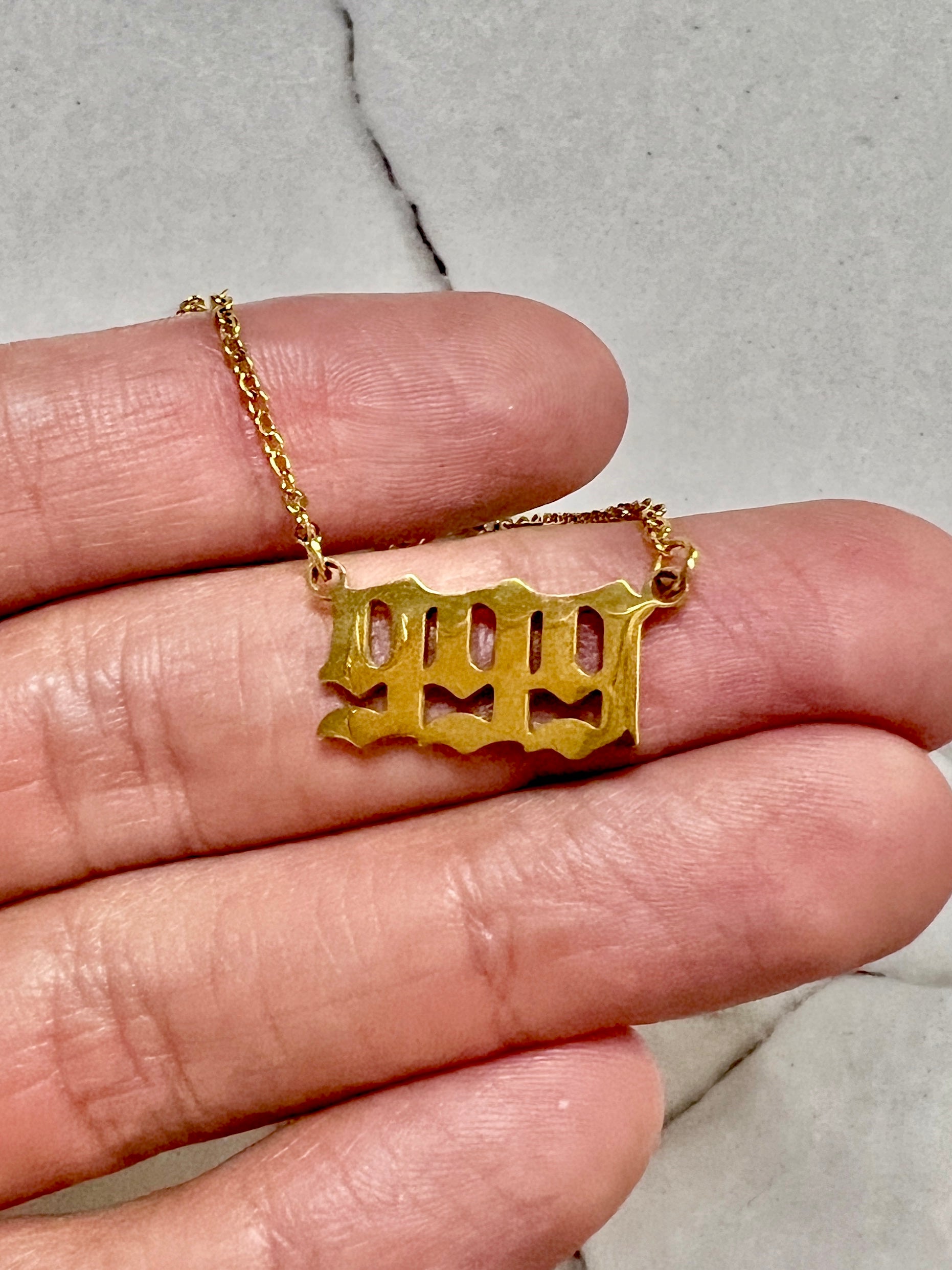 A gold-toned necklace with a stylized cutout 999 number pendant is draped over a person's fingers against a light gray marble background.