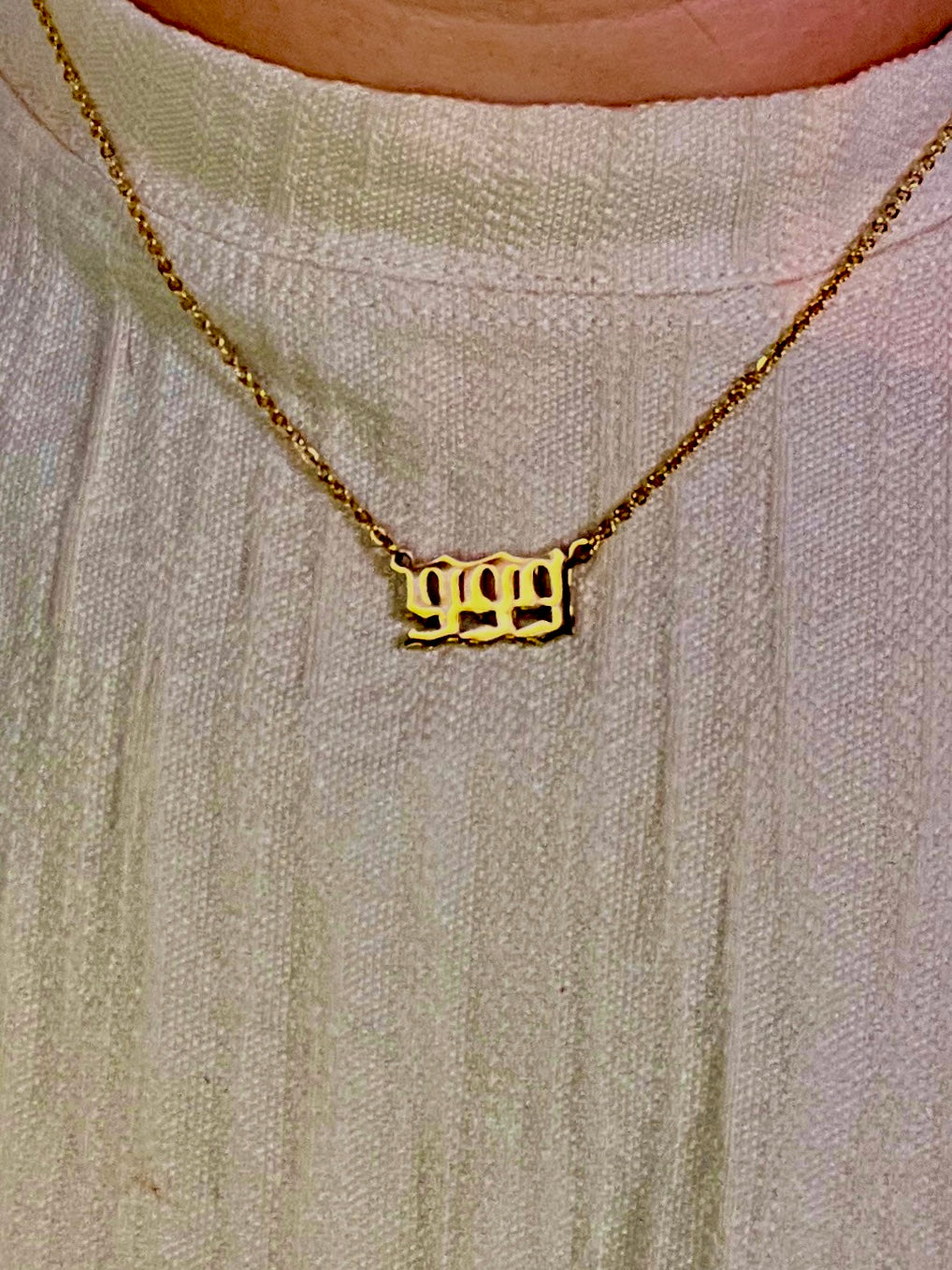 A close-up of a gold chain necklace with a custom 999  number  pendant spelling '999', worn by a person wearing a textured off-white top