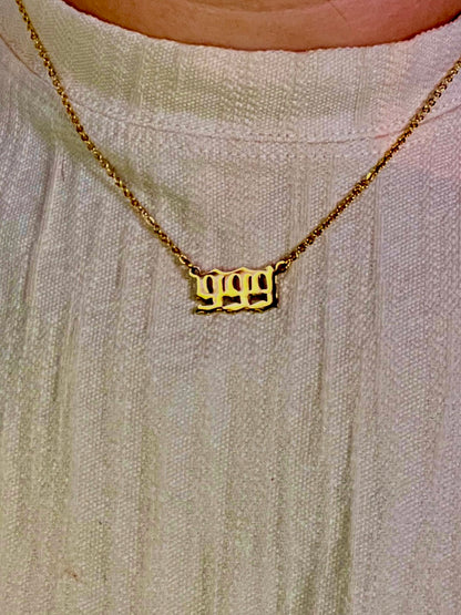 A close-up of a gold chain necklace with a custom 999  number  pendant spelling '999', worn by a person wearing a textured off-white top