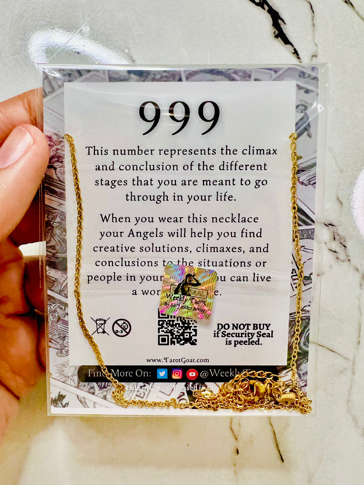 A gold necklace with a clear security seal charm is displayed on its retail backing card, held by a hand over a white marble surface. The card features a white background with black text detailing the '999' number meaning and a small illustration of a winged angel.