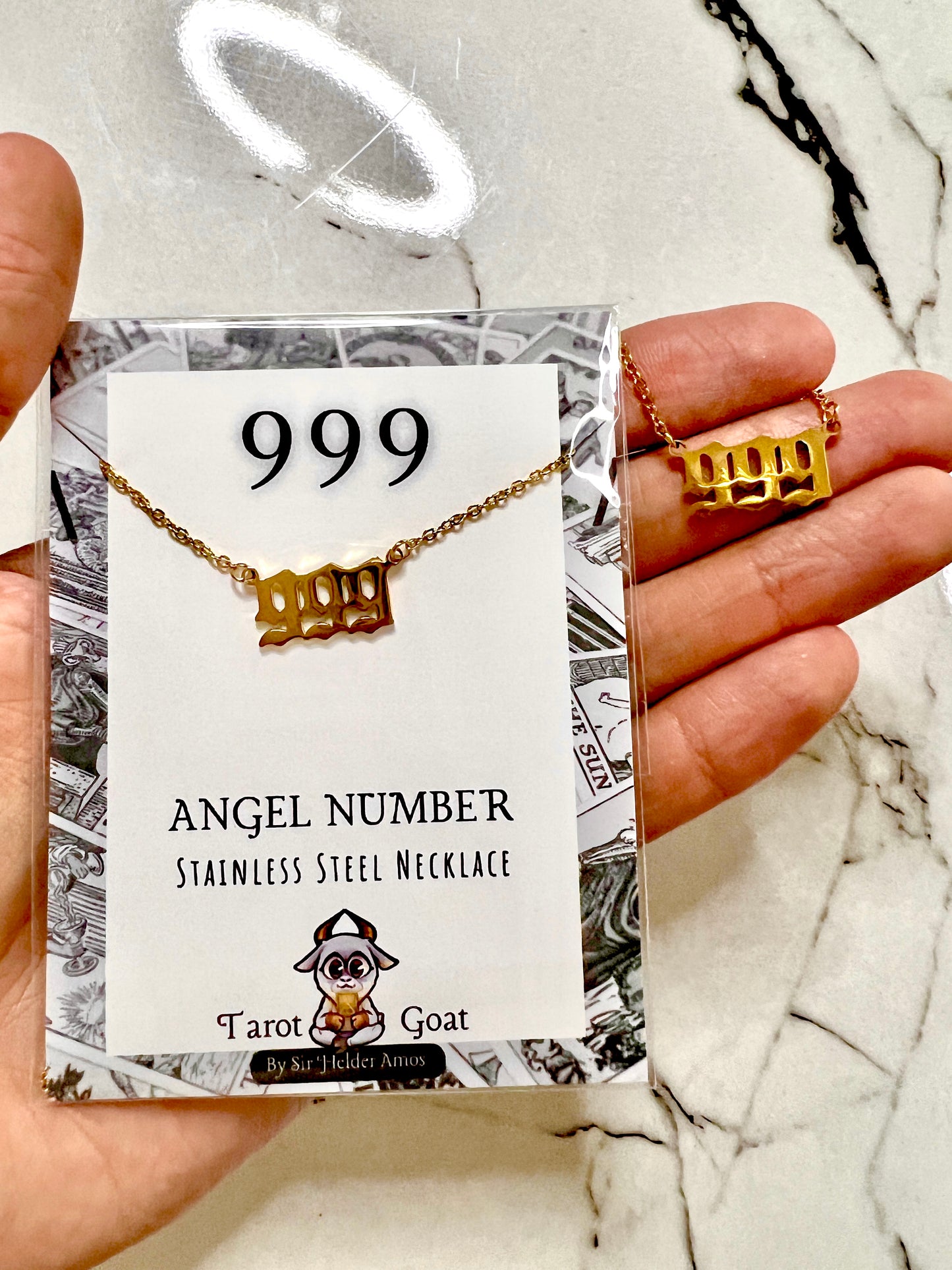A hand holds a gold-tone stainless steel necklace shaped like the number 999, displayed on a branded card labeled '999 ANGEL NUMBER STAINLESS STEEL NECKLACE' with the brand 'Tarot Goat'.