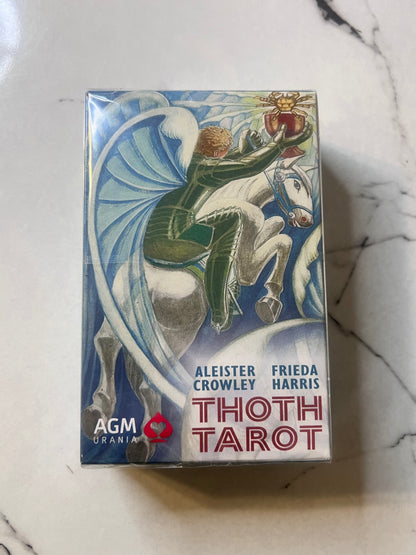 A boxed deck of Thoth Tarot cards by AGM Trania, featuring illustrated artwork of a rider on a white horse with blue wings, photographed on a light marble-like surface.