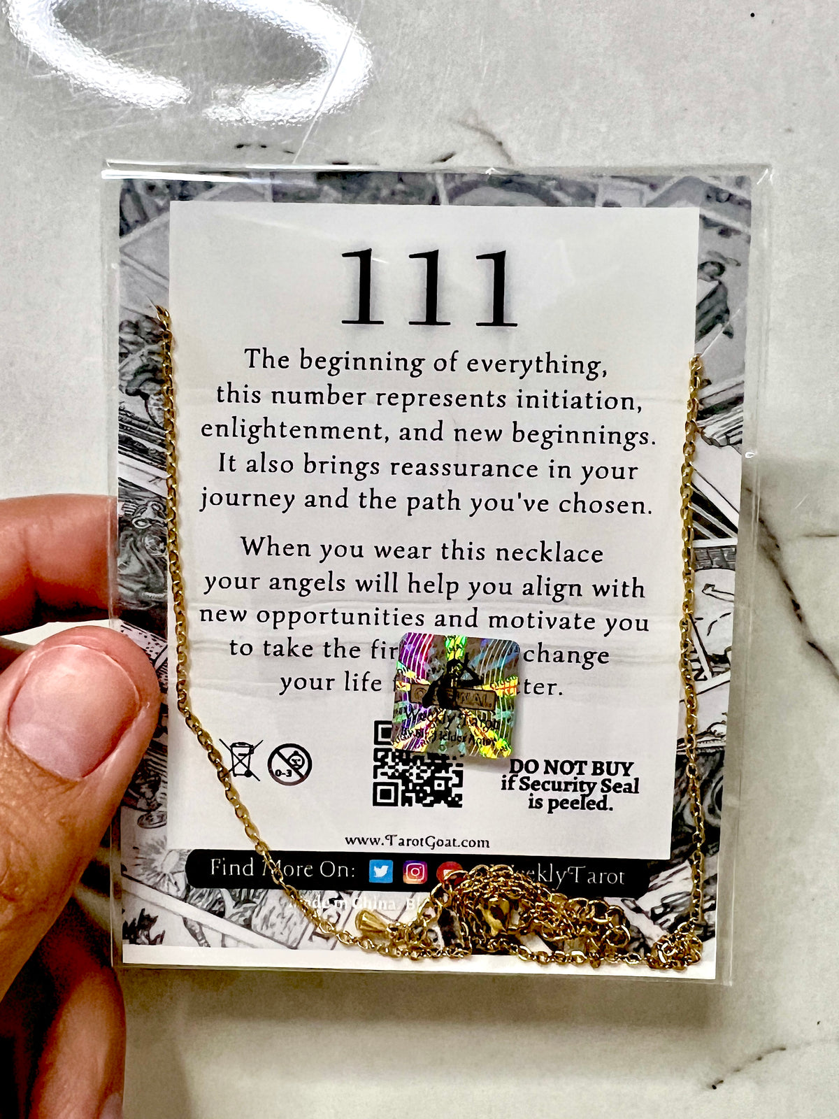A hand holding a clear plastic card containing a gold necklace with athe number 111  over a card that reads '111' and includes a tarot card reference. The background is a light gray marble-like surface.