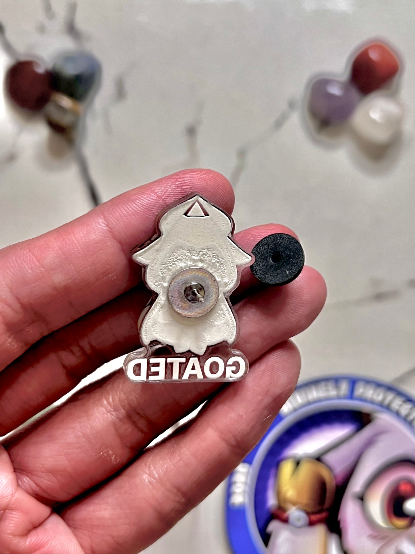 A hand holding a small silver metal pin shaped like a cartoon character with a black circular accent and the word 'GOATED' in white. The background is a light marble surface with blurred colorful beads and a circular container.