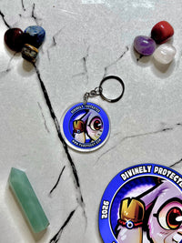A circular acrylic keychain with a cartoon goat wearing a red evil eye bracelet and the text 'DIVINELY PROTESTED' 
